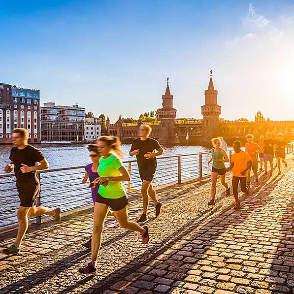 Im Detail zeigt das Bild eine Reihe von Menschen, die entlang eines gepflasterten Weges joggen, der an einem Fluss entlangführt. Im Hintergrund sind die Oberbaumbrücke und die Berliner Skyline zu sehen. Die Sonne scheint von der rechten Seite des Bildes her und scheint einen goldenen Schein auf die Szene zu werfen. Die Läufer sind unterschiedlich gekleidet, manche tragen Sportkleidung, andere eher alltägliche Kleidung. Sie scheinen in verschiedenen Stadien ihrer Laufrunde zu sein – einige laufen schneller, andere langsamer. Die Atmosphäre des Bildes ist aktiv und dynamisch, aber auch friedlich und entspannend. Die Stimmung ist positiv und optimistisch, vermittelt ein Gefühl von Gemeinschaft und gesundem Lebensstil. Die Steine des Weges scheinen alt und abgenutzt zu sein, was dem Bild einen Hauch von Geschichte und Tradition verleiht. Die Farben sind warm und einladend, mit dem Gold der untergehenden Sonne, die den Himmel und die Gebäude erleuchtet. Das Bild vermittelt ein Gefühl von Bewegung, Energie und der Schönheit Berlins.