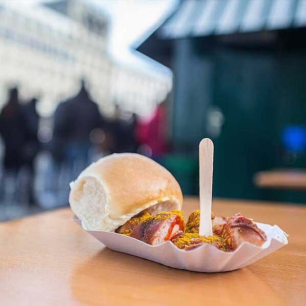 Das Bild zeigt eine Currywurst, die auf einem kleinen, weißen Pappteller liegt. Die Currywurst besteht aus mehreren mit Currypulver und Ketchup beträufelten Würstchenstücken. Ein weiches, helles Brötchen liegt neben den Würstchen auf dem Teller. Ein kleiner, hölzerner Spieß steht aufrecht in der Mitte der Currywurst.
Der Hintergrund ist unscharf, aber man kann erkennen, dass sich das Bild an einem belebten Ort in einer Stadt abspielt. Im Hintergrund sind Gebäude und Menschen, die sich bewegen oder an Tischen sitzen, zu sehen, was auf einen Markt oder eine belebte Straße hindeutet. Die Atmosphäre wirkt lebhaft und urban, mit einer leicht verschwommenen, aber dennoch klaren Darstellung der Umgebung. Die Farben sind warm und einladend, mit den goldenen Tönen des Currypulvers, dem roten Ketchup und dem hellen Brötchen, die einen Kontrast zum etwas kühleren, aber dennoch freundlichen Hintergrund bilden. Die gesamte Stimmung ist gemütlich und appetitanregend, die Currywurst steht im Fokus und wirkt als ein köstlicher Imbiss.