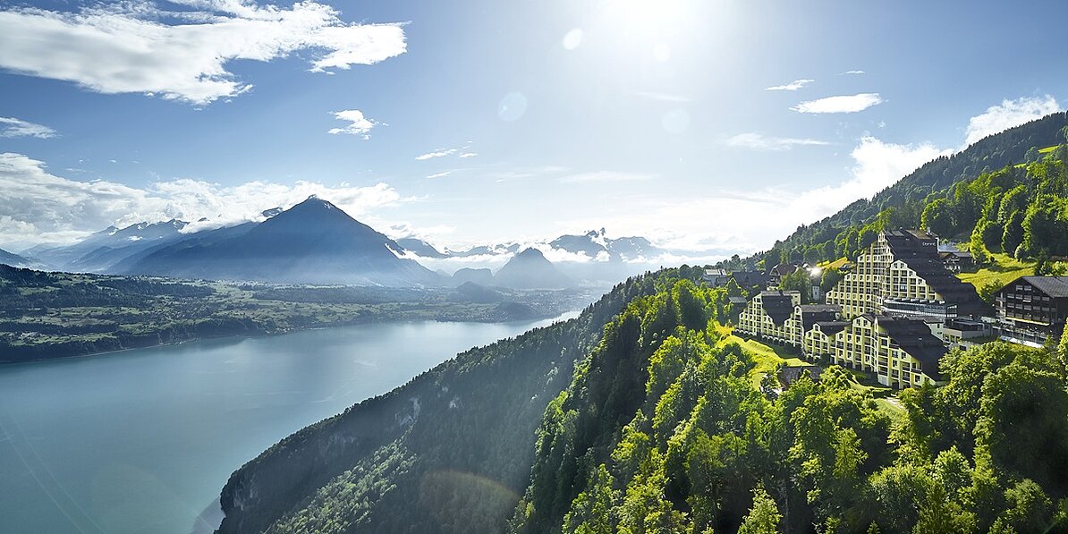 Dorint Hotel Blüemlisalp Beatenberg on a forested mountainside with panoramic views of Lake Thun and the Alpine mountain landscape in the background.