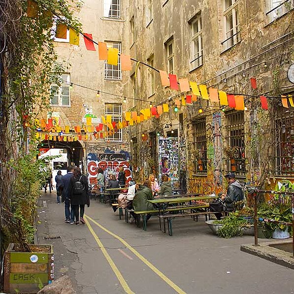 Im Detail zeigt das Bild eine schmale Gasse zwischen alten, verwitterten Gebäuden, die mit Graffiti und Plakaten verziert sind. Bunte Fahnen in Orange und Gelb hängen über die Gasse gespannt und schaffen ein Festliches Ambiente. Mehrere Picknicktische und Bänke stehen im Hof, an denen sich Menschen unterhalten und entspannen. Einige Personen gehen durch den Hof, während andere an den Tischen sitzen. Die Kleidung der Menschen ist alltäglich und ungezwungen. Die Gebäude wirken alt und etwas heruntergekommen, aber sie sind mit Pflanzen und Graffiti lebendig gestaltet. Die Stimmung ist entspannt, freundlich und lebendig, fast schon ein bisschen bohemmisch. Ein kleiner, begrünter Bereich ist ebenfalls zu sehen, der zu der natürlichen Atmosphäre beiträgt. Im Hintergrund ist ein Tor oder Durchgang zu sehen, der auf einen weiteren Bereich hindeutet. Insgesamt herrscht eine fröhliche und einladende Atmosphäre, die auf einen gemütlichen Treffpunkt oder ein Café hindeutet.