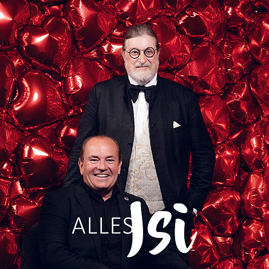 Das Bild zeigt zwei Männer in Abendgarderobe vor einem Hintergrund aus roten Luftballons in Herzform; es handelt sich um ein Werbefoto für den Podcast „Alles Isi“.