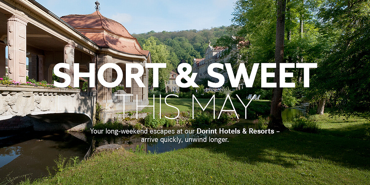 The words “Short & Sweet this May” in the foreground, with a pavilion, a green meadow, and water in the background