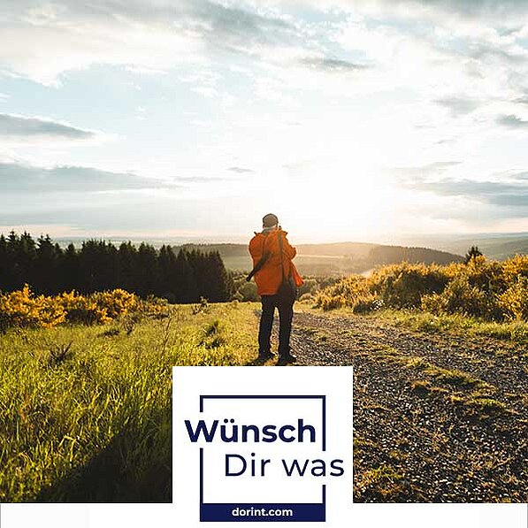 Das Bild zeigt einen Mann mit einer orangenen Jacke und einer schwarzen Hose, der einem Feldweg entlang geht, mit dem Rücken zur Kamera. Er trägt einen Kameragurt über der Schulter und scheint eine Kamera zu halten, während er in die Ferne blickt. Die Landschaft um ihn herum ist malerisch, mit hoch aufragenden Bäumen und Hügeln in der Ferne, die von einem goldenen Sonnenlicht beleuchtet werden. Das Licht lässt die Szene warm und einladend wirken, während der Himmel einen Hauch von Wolken zeigt, die zur allgemeinen Idylle beitragen. Der Mann steht auf einem kleinen, unbefestigten Weg, der durch einen grünen und gelben Bereich verläuft, der mit hohem Gras und niedrigen Büschen bedeckt ist. Im Vordergrund dominieren die langen Gräser, die im Sonnenlicht glänzen. Die Atmosphäre ist ruhig und friedlich, und das Bild vermittelt ein Gefühl von Ruhe und dem Abenteuer in der Natur. Das Bild enthält auch eine weiße Box mit der Aufschrift "Wünsch Dir was" und einer Website-URL darunter, was auf eine Werbung oder eine Kampagne hindeutet.