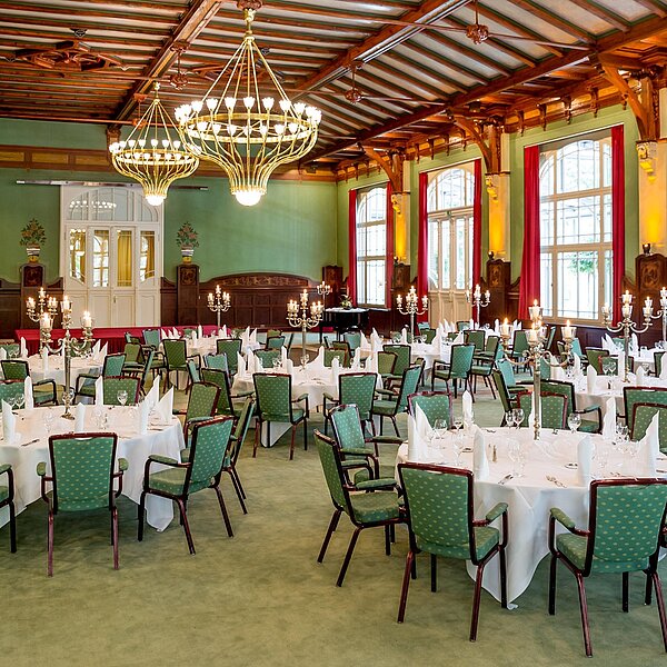 Festlich gestalteter Jugendstilsaal mit Kronleuchtern, dunklem Holz, grünem Teppichboden und elegant gedeckten Tischen.