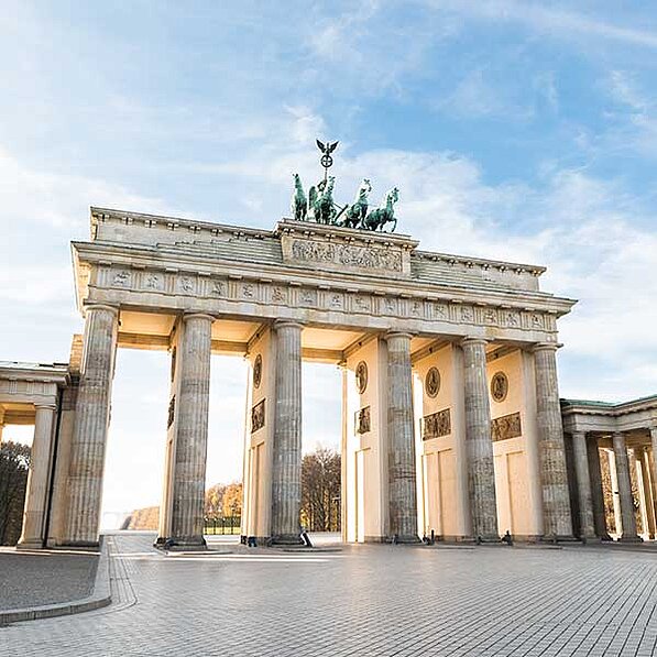 Das Bild zeigt eine weitläufige Aufnahme des Brandenburger Tors in Berlin, Deutschland. Das Tor, ein neoklassizistisches Bauwerk aus Sandstein, steht im Zentrum des Bildes und ist von einem großzügig angelegten Platz umgeben. Auf dem Tor selbst sind mehrere Säulen zu sehen, die einen großen Triumphbogen bilden. Oben auf dem Tor befindet sich eine Quadriga, eine von vier Pferden gezogene Wagen, auf der sich eine geflügelte Siegessäule befindet. Die Quadriga ist aus Bronze und detailliert gestaltet.
Die Säulen des Tores sind hoch und majestätisch und tragen zur imposanten Erscheinung des Bauwerks bei. Der Sandstein des Tores erscheint hell und warm im Sonnenlicht. Der Platz vor dem Tor ist weitläufig und gepflastert, im Hintergrund erkennt man Bäume und ein Stück des Himmels. Der Himmel ist hellblau mit wenigen Wolken, was auf einen sonnigen und klaren Tag hindeutet.
Die gesamte Szenerie wirkt ruhig und friedlich. Die Lichtverhältnisse und die Architektur schaffen eine Atmosphäre von Würde und Geschichte. Das Fehlen von vielen Menschen verstärkt die Ruhe und erlaubt es dem Betrachter, sich auf die Architektur und Monumentalität des Brandenburger Tores zu konzentrieren. Das Bild vermittelt einen Eindruck von Größe und Bedeutung des Bauwerks als historisches und kulturelles Symbol Berlins und Deutschlands.