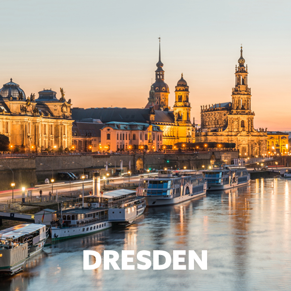 Das Bild zeigt die atemberaubende Skyline von Dresden bei Sonnenuntergang, mit beleuchteten historischen Gebäuden entlang der Elbe und mehreren Ausflugsschiffen, die an den Ufern festgemacht haben.