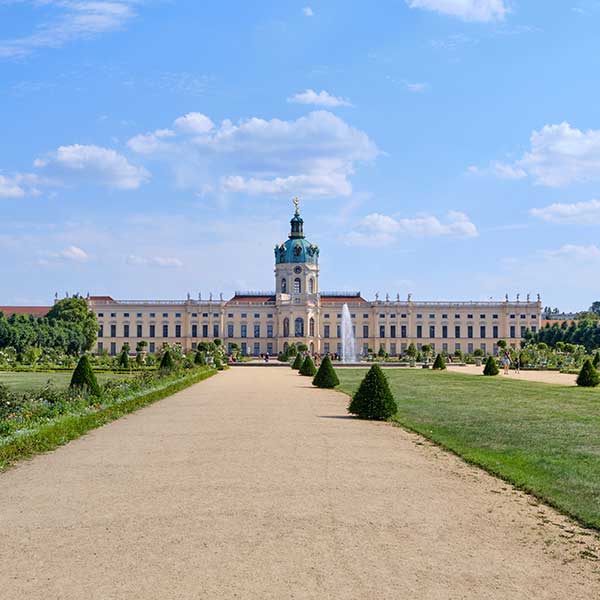 Das Bild zeigt das Schloss Charlottenburg in Berlin an einem sonnigen Tag von einem langen, sandigen Weg aus, der von formalen Gärten gesäumt ist.