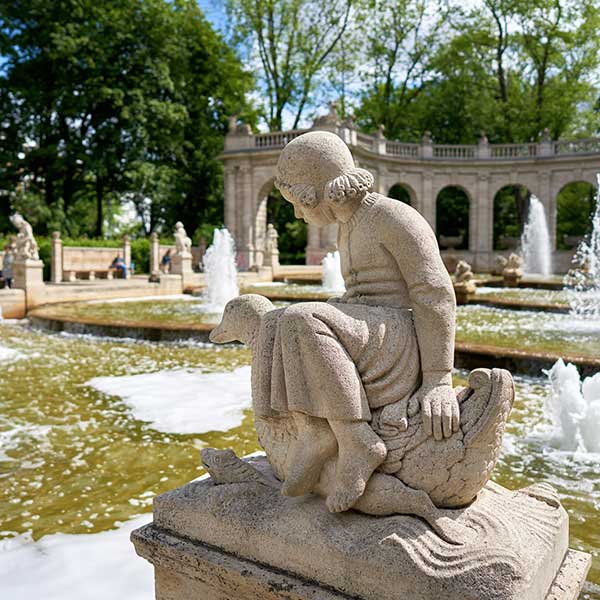 Das Bild zeigt eine Steinstatue eines Kindes, das auf einem Vogel sitzt, vor dem Hintergrund eines prächtigen Brunnens in einem Park.