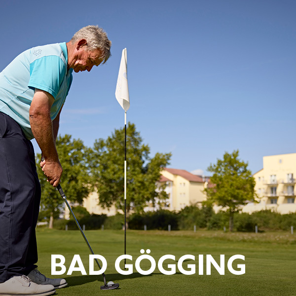 Mann beim Golfabschlag auf den Grün. Im Hintergrund eine Fahne und die Front der Hotelanlage des Dorint Marc Aurel Resort Bad Gögging