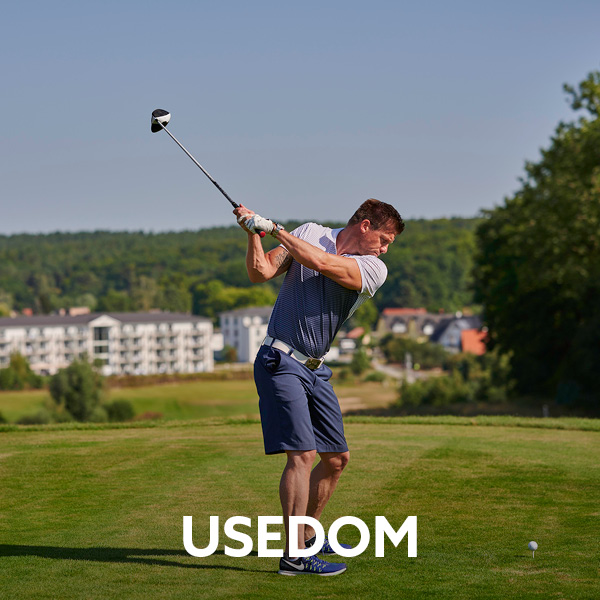 Mann holt aus zum Golfabschlag vor ihm der Ball aufgeteet auf dem Grün. Im Hintergrund die helle Hotelanlage des Dorint Resort Baltic Hills Usedom